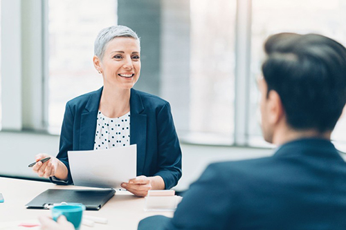 Smiling person with short gray hair, in a business suit, holds documents while talking to a person in an office setting, conveying a professional tone.