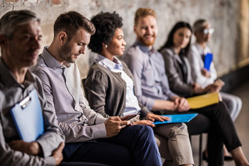 A diverse group of six people sit in a row against a brick wall, each holding folders, engaged and waiting, conveying a professional and anticipatory mood.