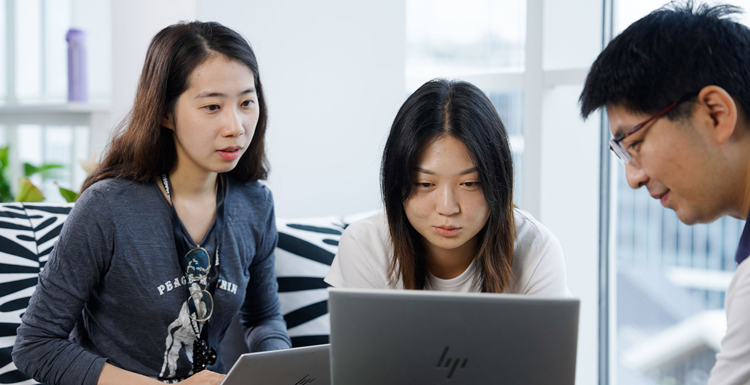 Three people sitting on a couch looking at a laptop in an office environment