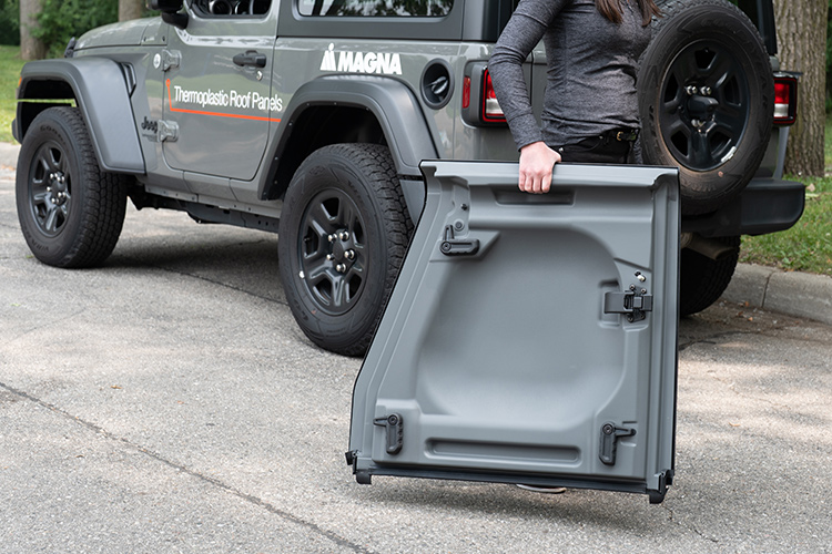 Person carrying thermoplastic roof panel with a Magna branded grey jeep in background