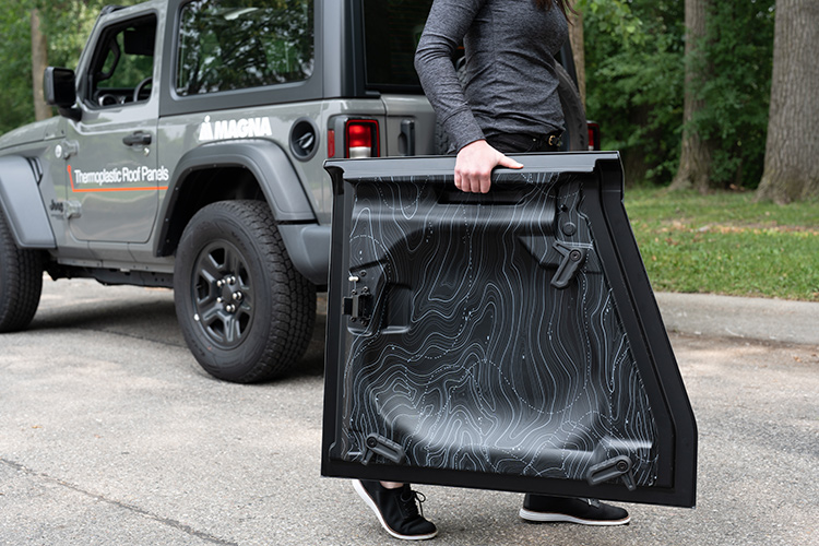 Person carrying thermoplastic roof panel with a Magna branded grey jeep in background