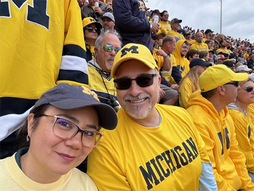 Jason Hegedus and other person smiling and wearing Michigan Wolverines jersey and sitting in the stands at Michigan Stadium