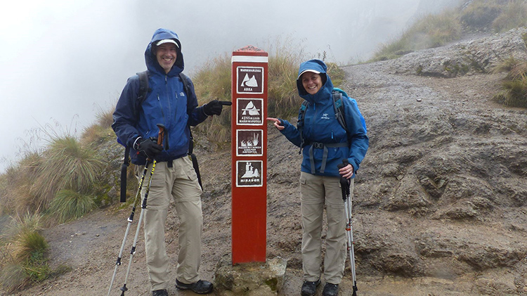 Maria Kennedy and another person dressed in blue jackets and khaki pants pointing at a sign as they are hiking up a mountain