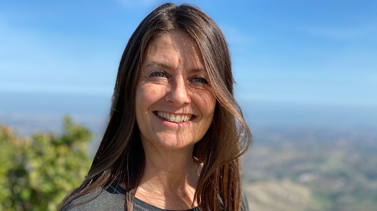 Maria Kennedy standing outside smiling with blue sky, trees and water in the background