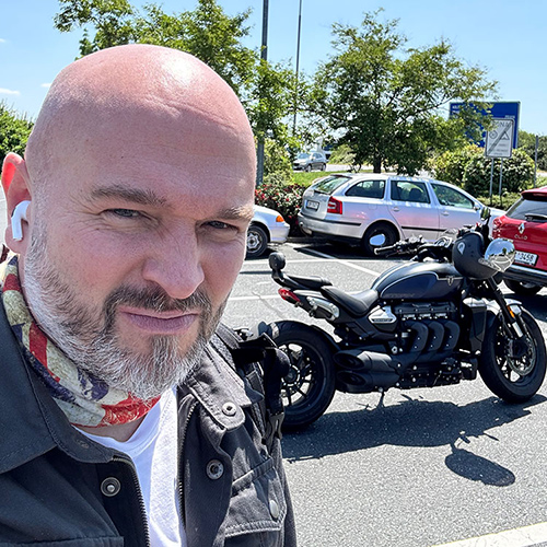 Miroslav Frontz with earphones and a scarf stands in a parking lot, smiling. A black motorcycle and cars are visible on a sunny day with trees in the background.